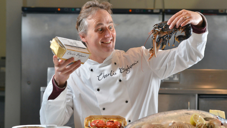 Charlie Bigham in a chef uniform with a large Charlie Bigham’s logo, holding a lobster in his left hand and a product in the right.