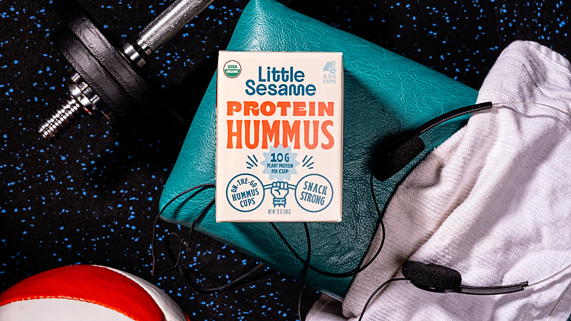 Little Sesame Protein Hummus ready-to-eat snack laid on top of a bag surrounded by a dumbbell, basketball, headphones and a towel.