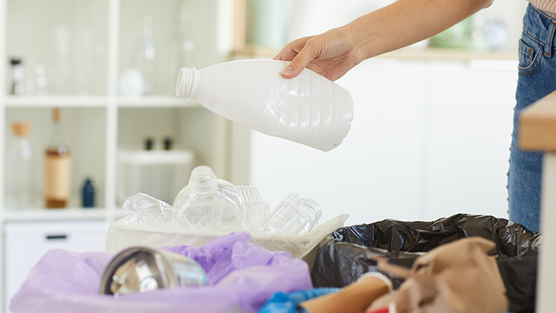 Close-up of a person recycling a white plastic bottle into the recycling bin. The blurred background is a white shelves with various bottles.