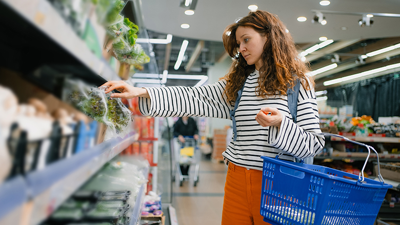Young woman looking at packaged greens on the shelf in a grocery store.  She is carrying a blue shopping basket.