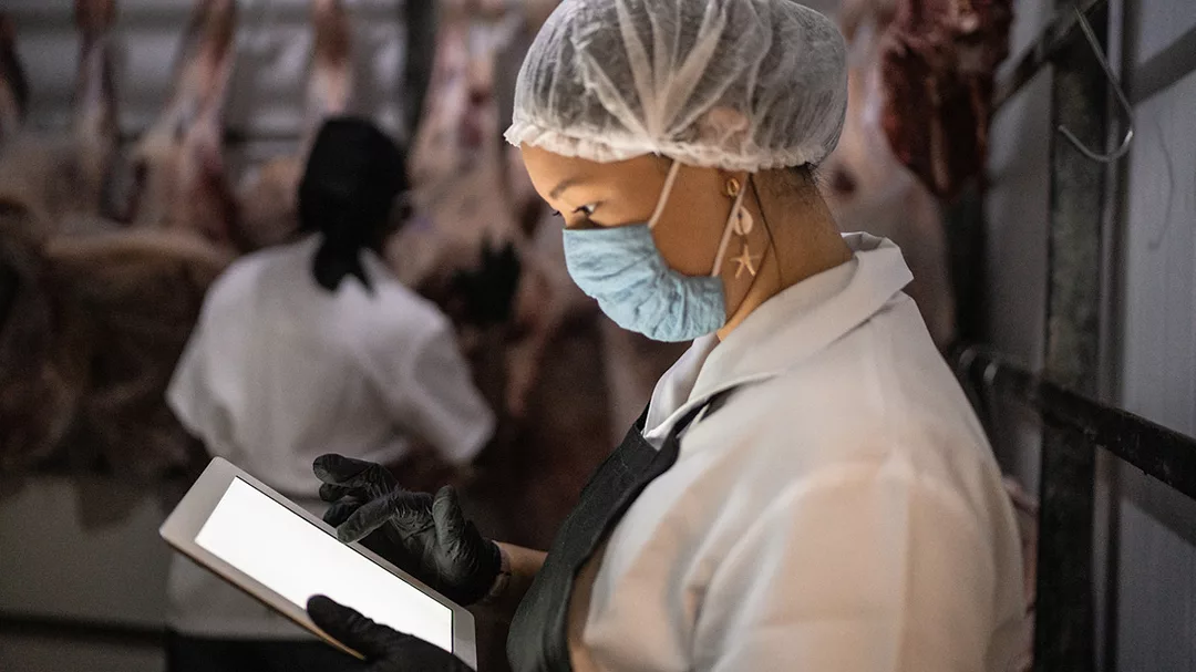 Female butcher using digital tablet with face mask at cold storage. Another worker is shown in the blurred background.