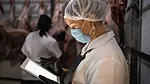 Female butcher using digital tablet with face mask at cold storage. Another worker is shown in the blurred background.