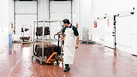 A male butcher is moving sausages on a pallet jack in a food processing plant.