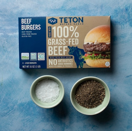 An overhead view of a package of Teton Waters Ranch Beef Burgers with two small bowls - left filled with salt, right filled with pepper - on a blue patterned table.