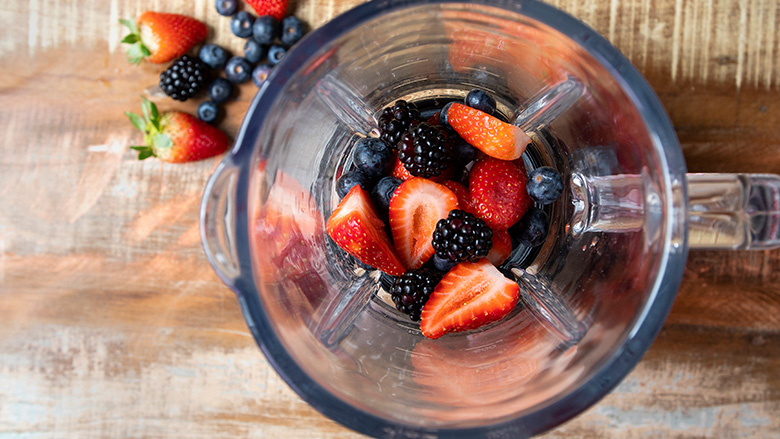 A overview shot of strawberries, blackberries and blue berries inside a blender. Some fruit in on the table.