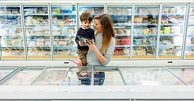 A mother is carrying her son and both are holding a frozen packaged item in the frozen section of a grocery store.