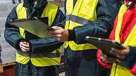 Close up and cropped image of three warehouse workers in safety gear talking to each other, two looking at a clipboard and one (far right) using a tablet.